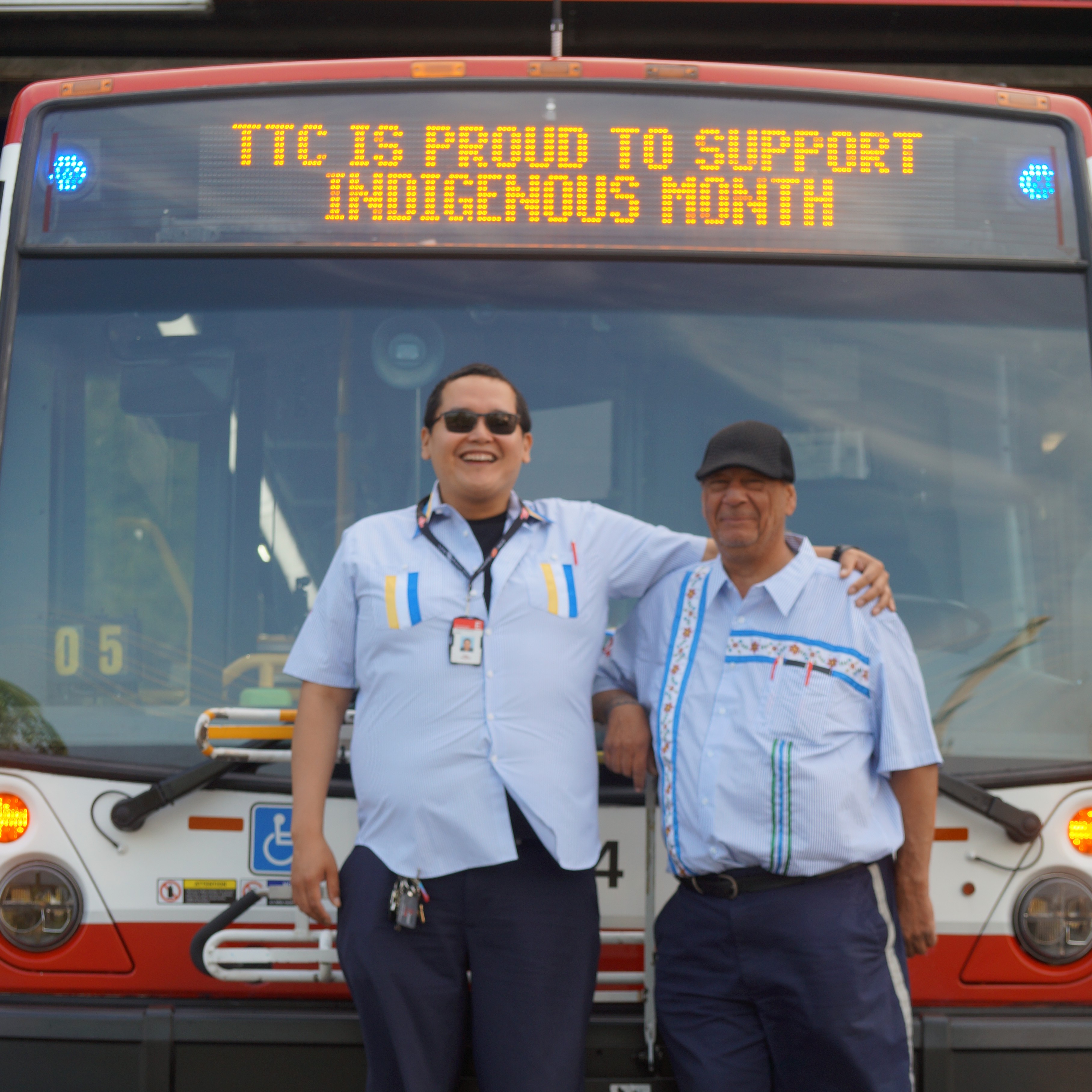 Two Indigenous TTC employees stand in front of a TTC bus with TTC is proud to support Indigenous Month displayed on the front of the bus. 