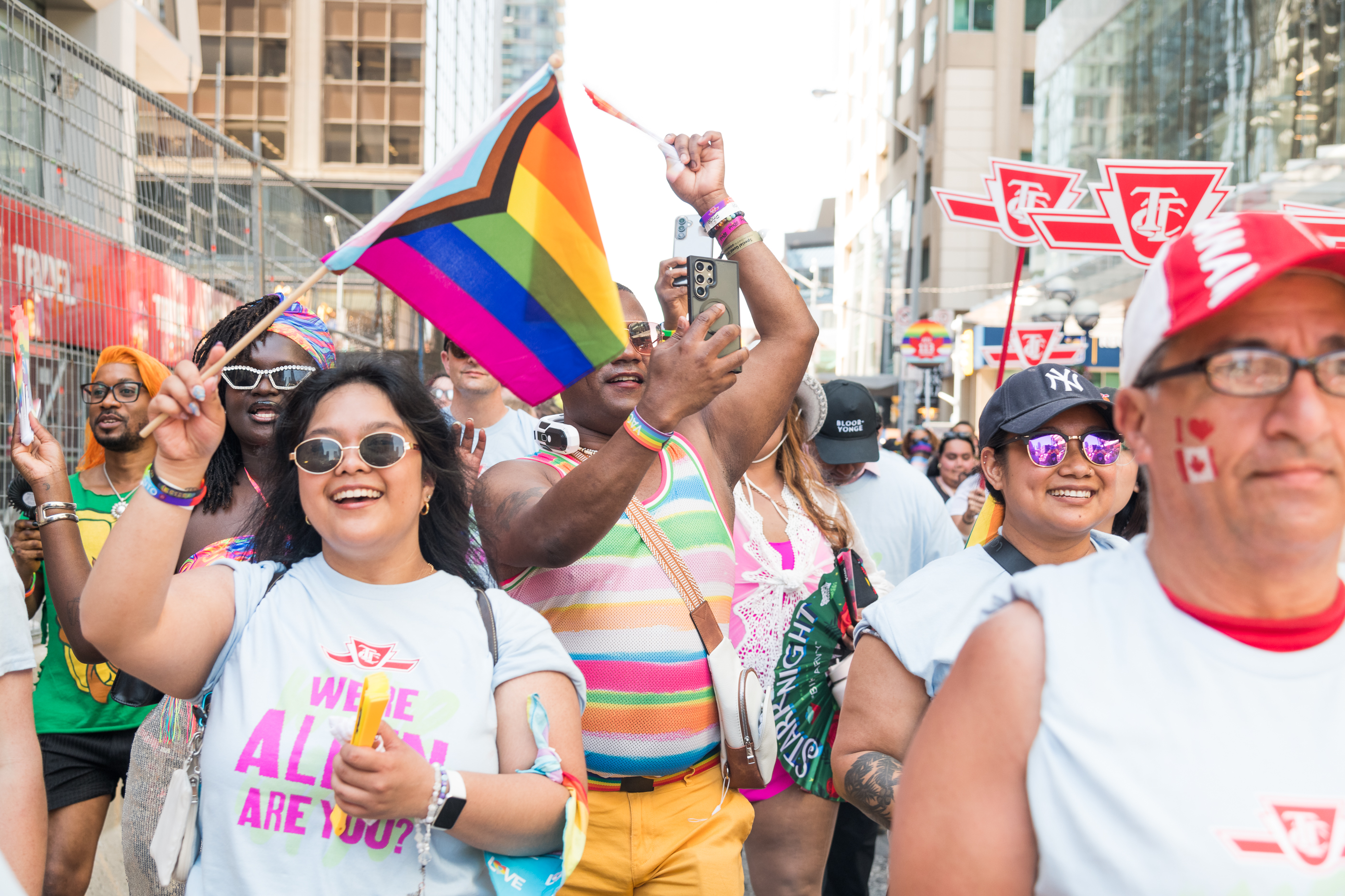 Group of people marching and smiling in the Toronto Pride Parade. One person at the front is holding a Progress Flag, and some people are holding red and white TTC signs.