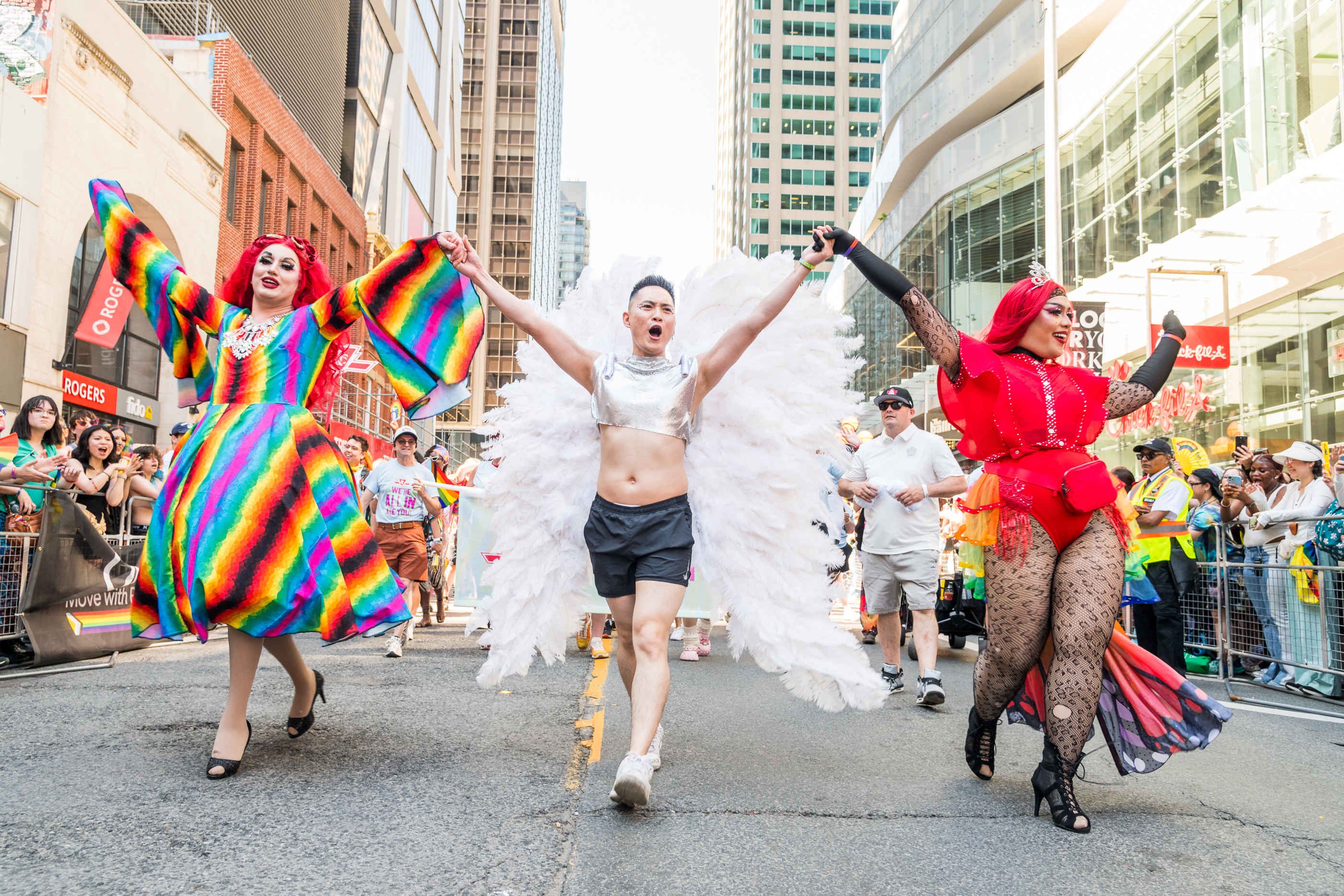 A person wearing large, white angel wings walks down the street as part of a Pride Parade. On either side of the person, there is a colourfully-dressed drag queen and they all have their arms raised.