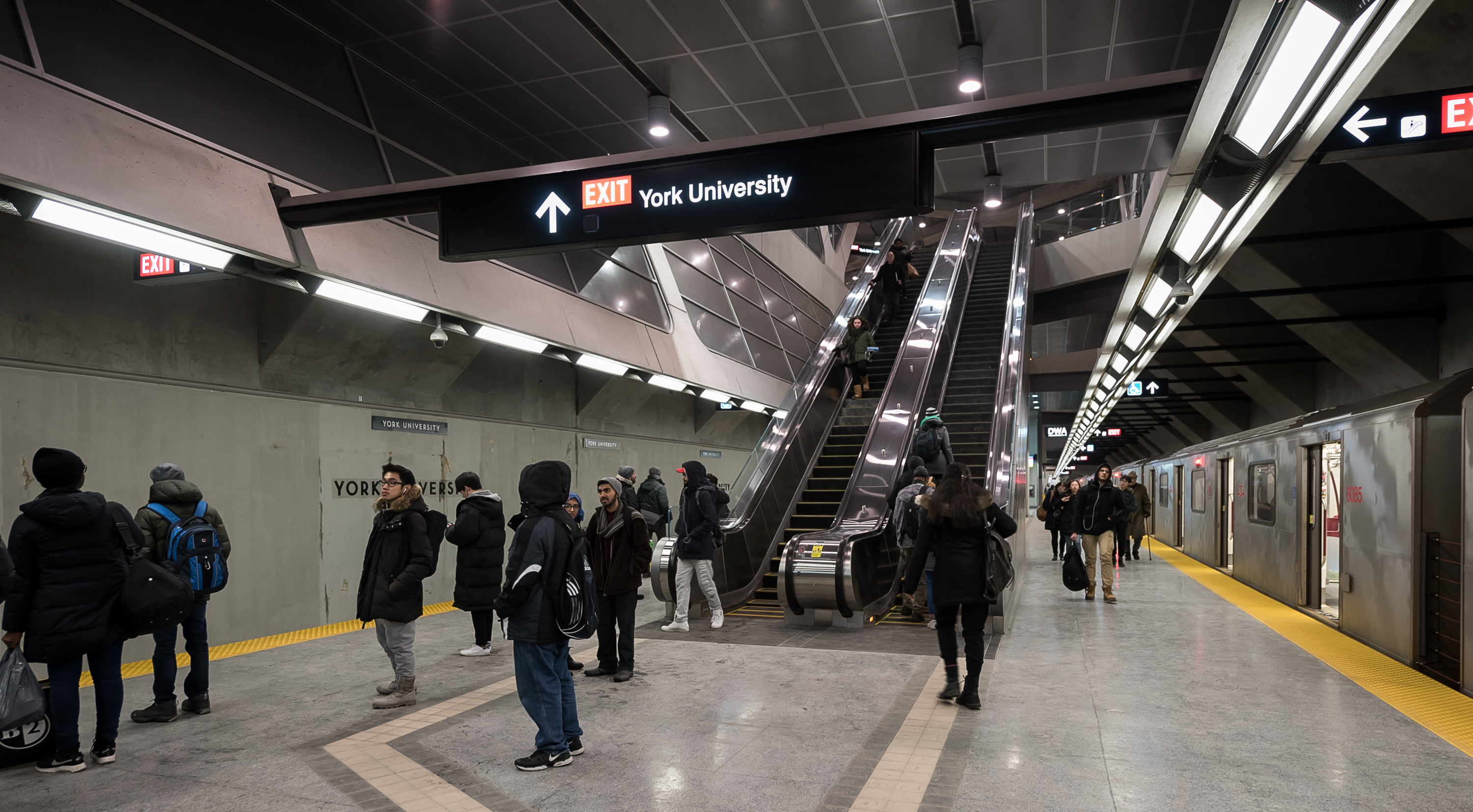 A group of people standing on a TTC subway station platform under a sign for York University. There is a train with open doors on one of the tracks.
