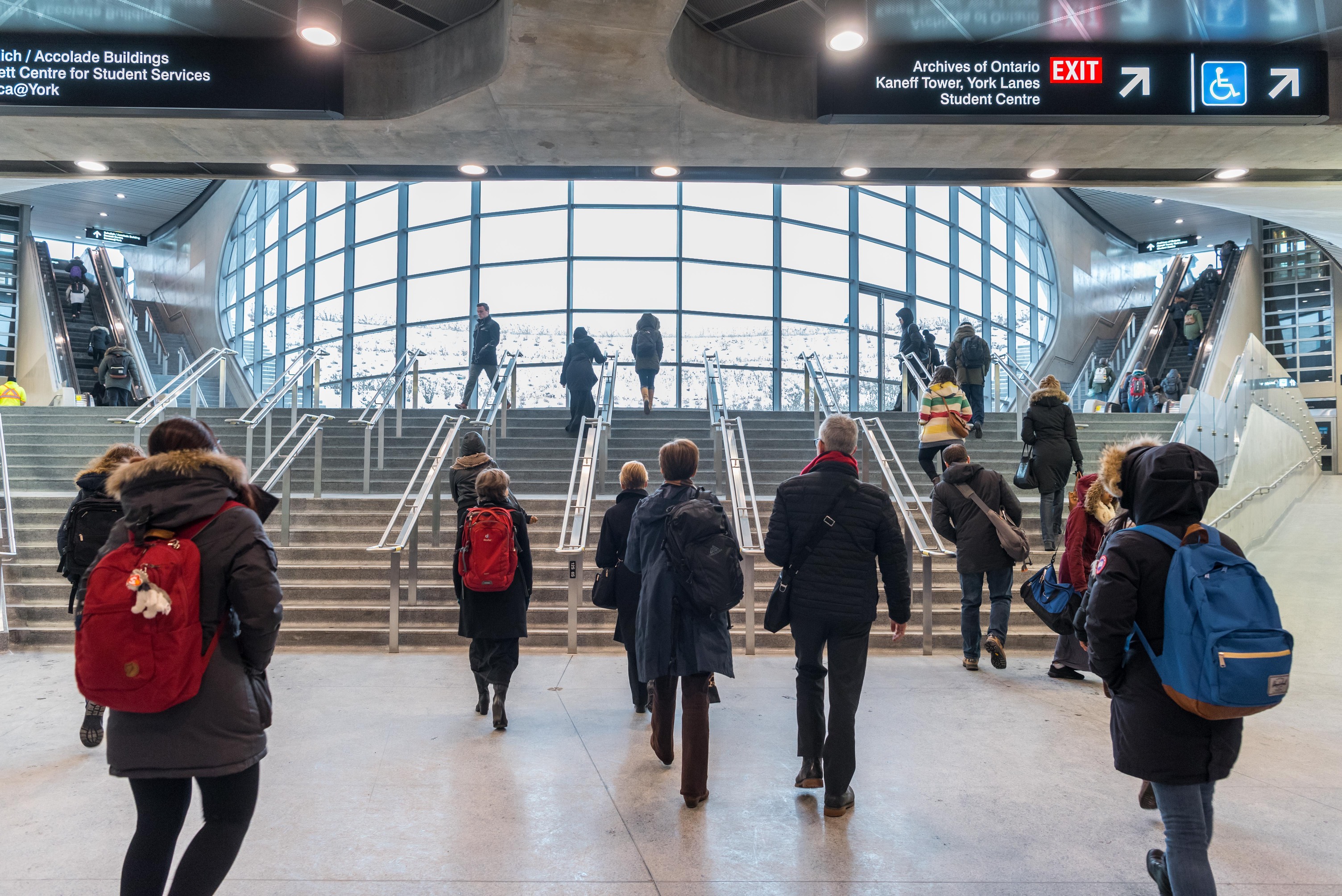 A group of people, many wearing backpacks, leaving a station via stairs and escalators.