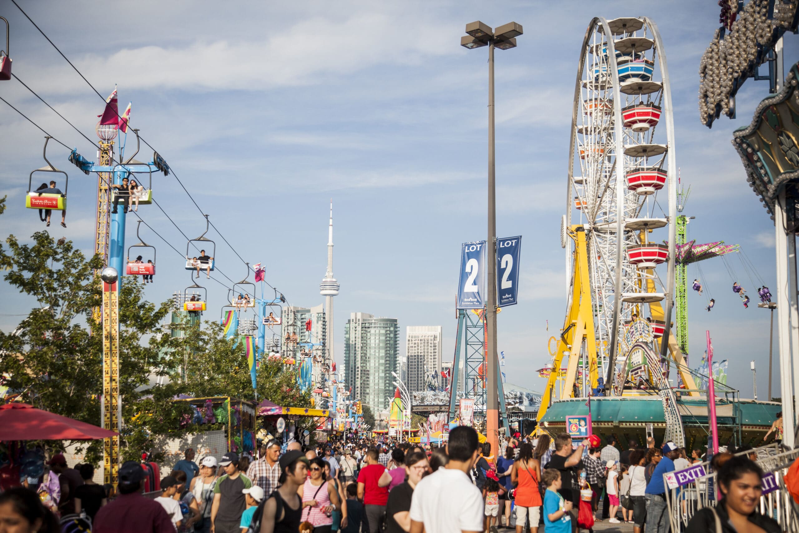 A crowd of people beside an open-air chairlift and a Ferris wheel at the Canadian National Exhibition.