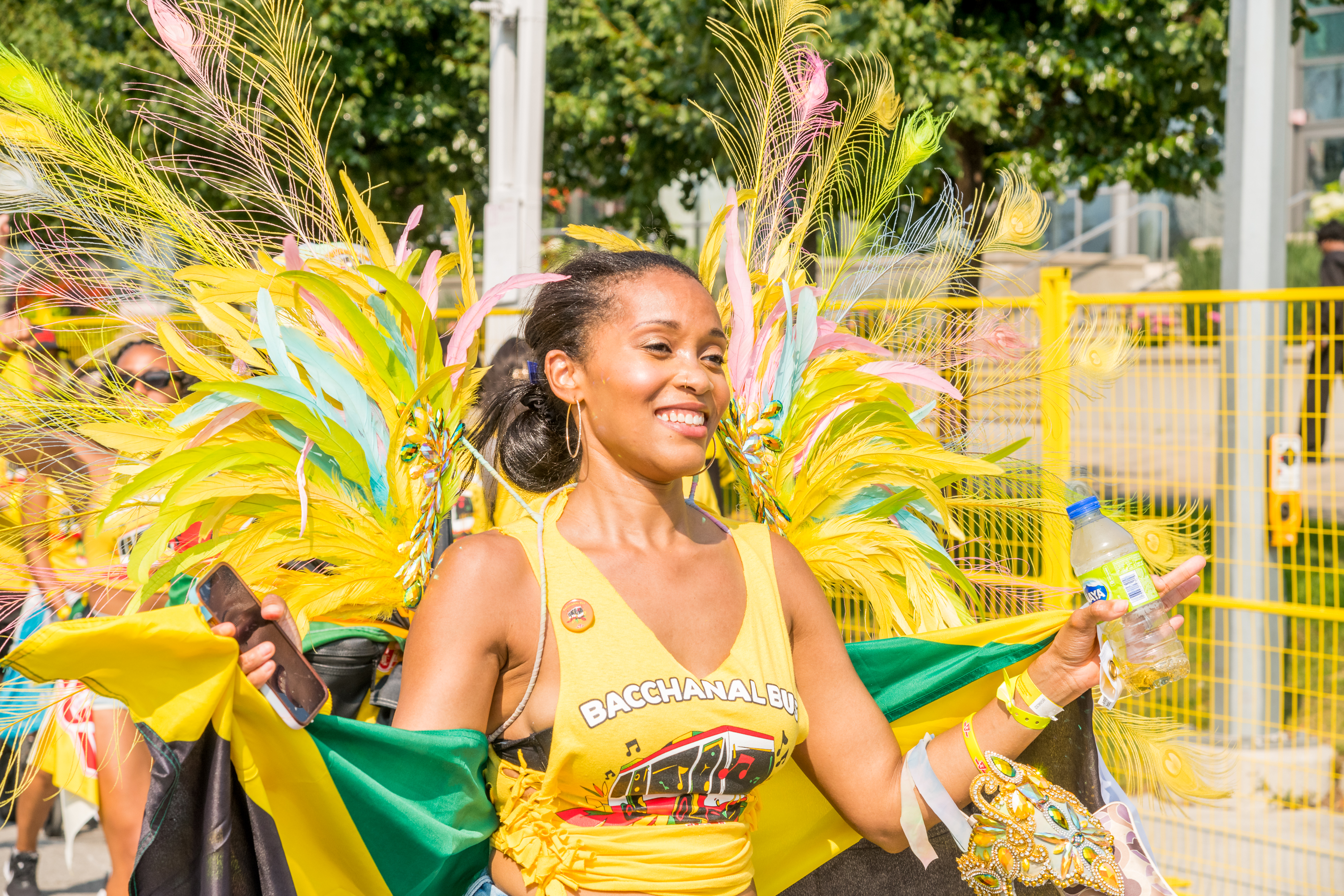 A person wearing a yellow TTC Bacchanal Bus shirt with feathers behind them walking down a street in the Toronto Caribbean Carnival Grand Parade.