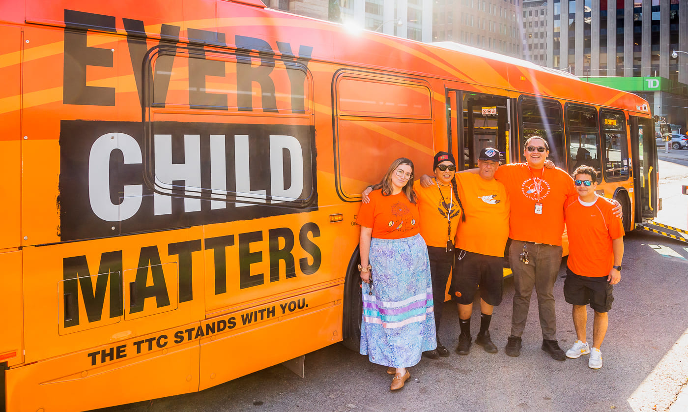 TTC staff wearing orange shirts stand beside an orange wrapped Every Child Matters TTC bus.