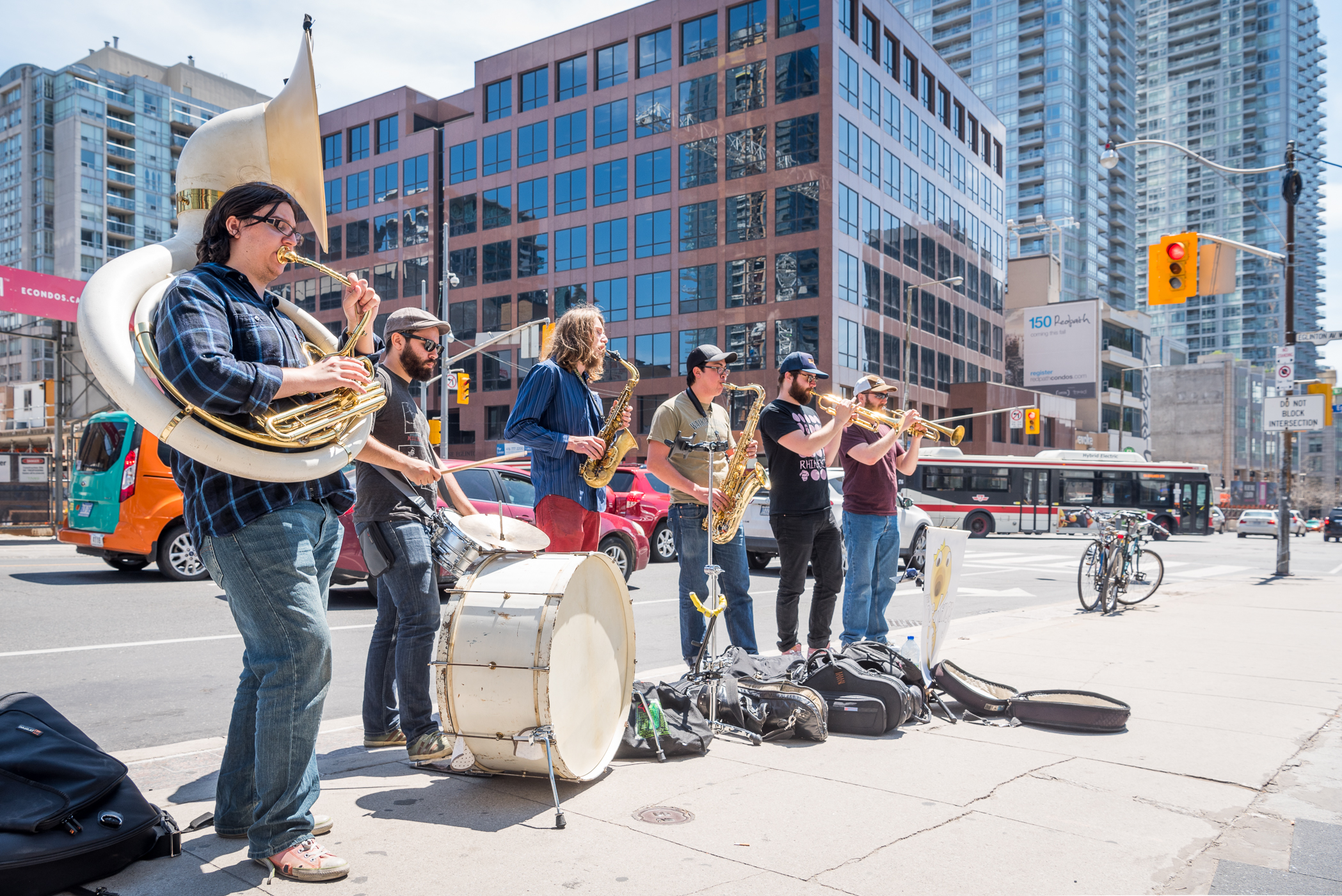Six musicians playing various instruments on a downtown Toronto street with a TTC bus in the background.