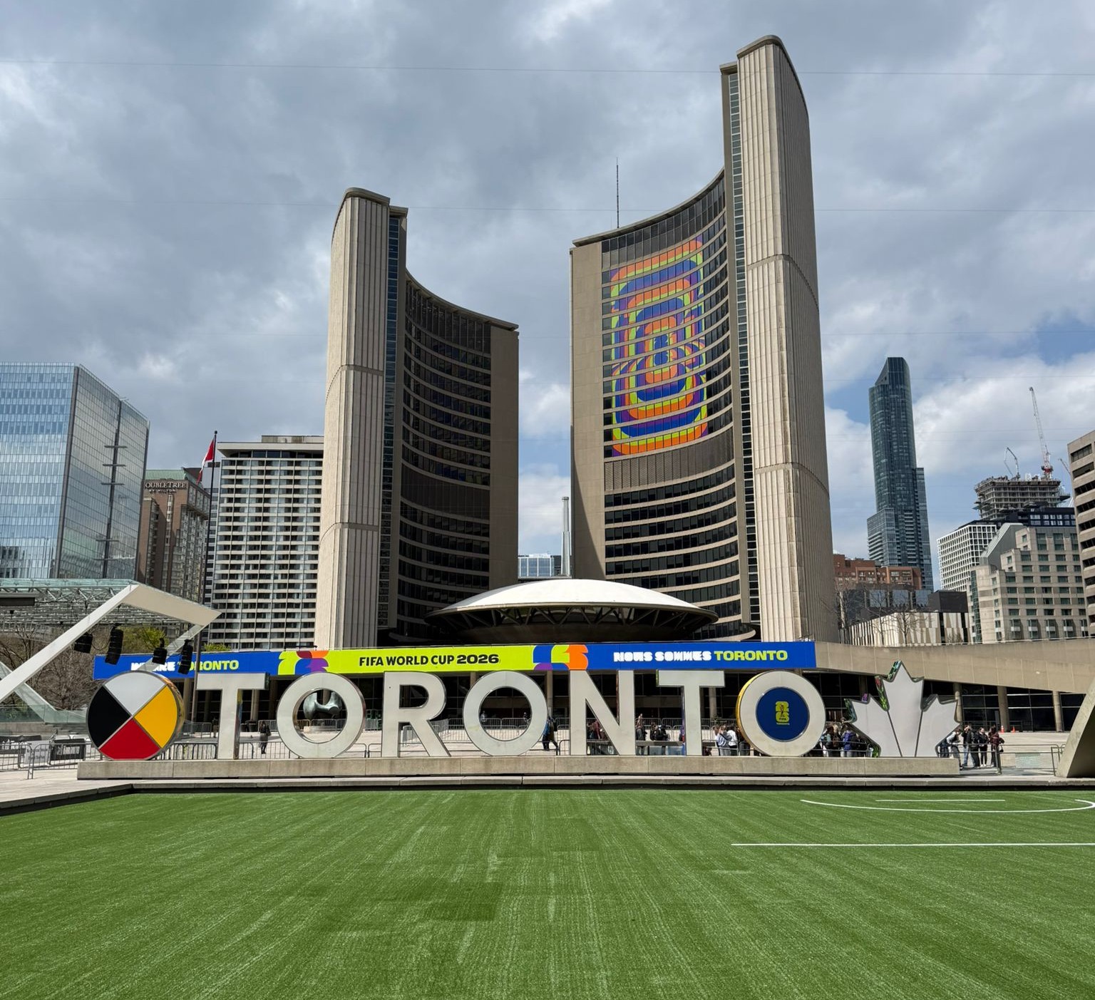 Nathan Phillips Square with a large Toronto sign, green soccer pitch and tall buildings in the background with a colourful logo.