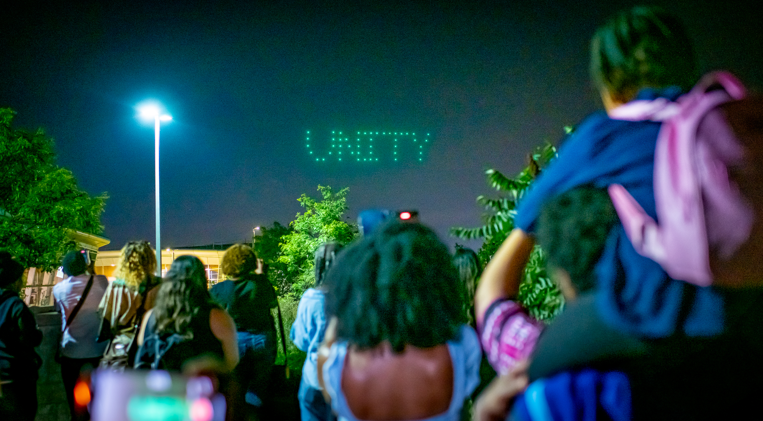 A large group of people from the back looking at a glowing green "unity" sign in the night sky.