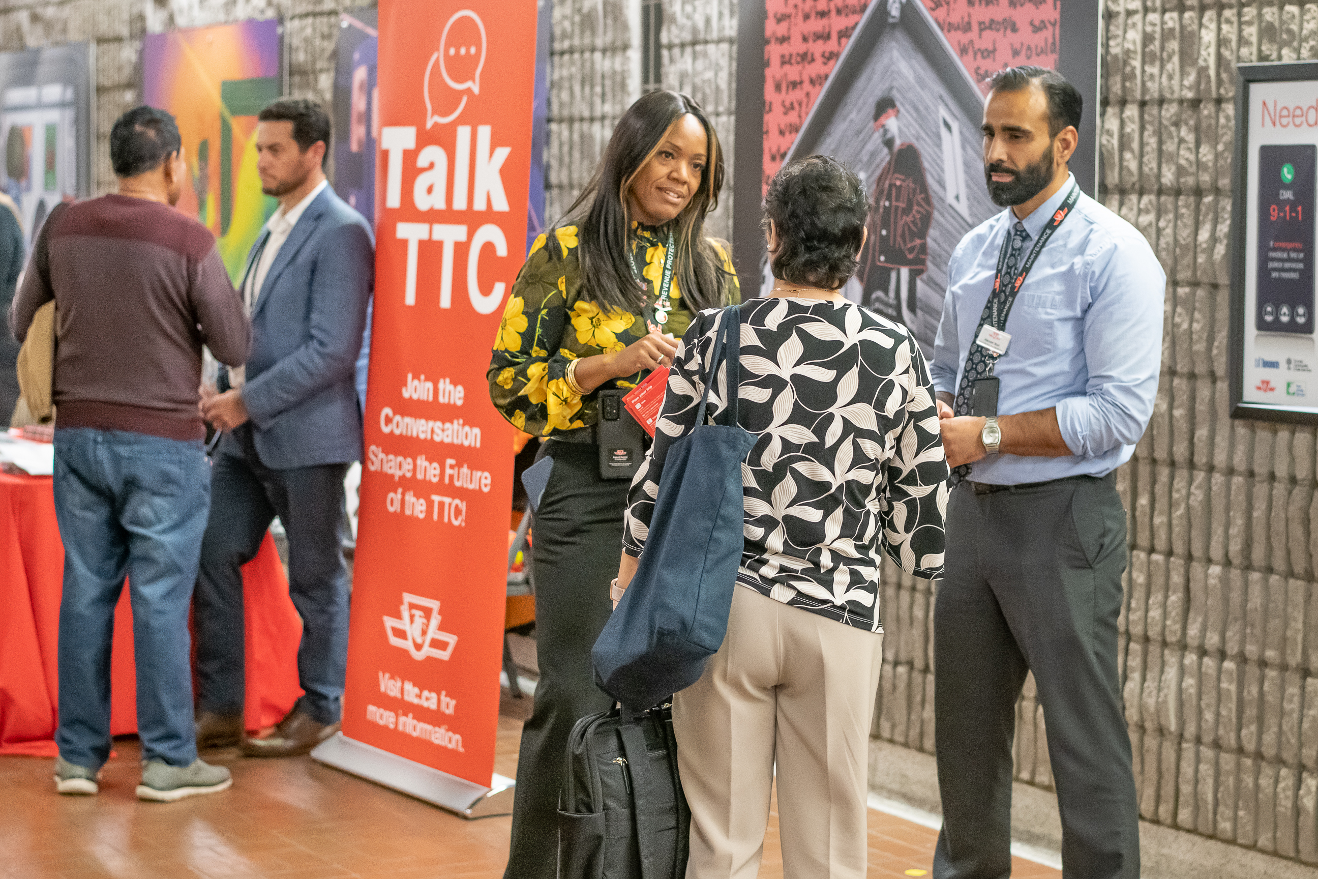 People engage in conversations at a public outreach event hosted by the Toronto Transit Commission (TTC). A bright orange banner with the words 'Talk TTC – Join the Conversation, Shape the Future of the TTC!' is prominently displayed. Two TTC staff members, wearing badges and lanyards, speak with attendees in a well-lit indoor space featuring promotional posters and informational displays.