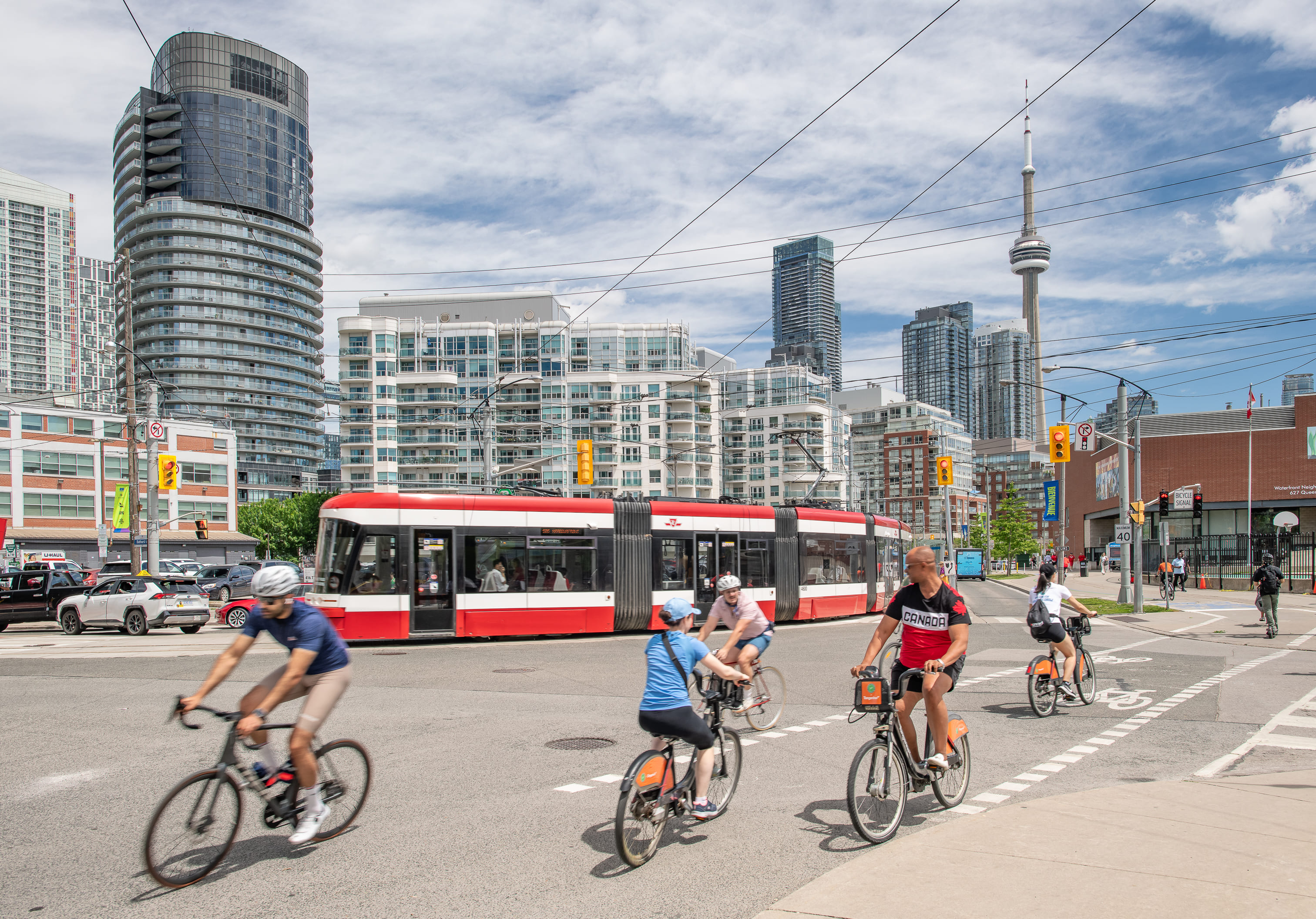 Cyclists crossing the street in front of a red and white TTC streetcar with condo buildings and the CN Tower in the background.