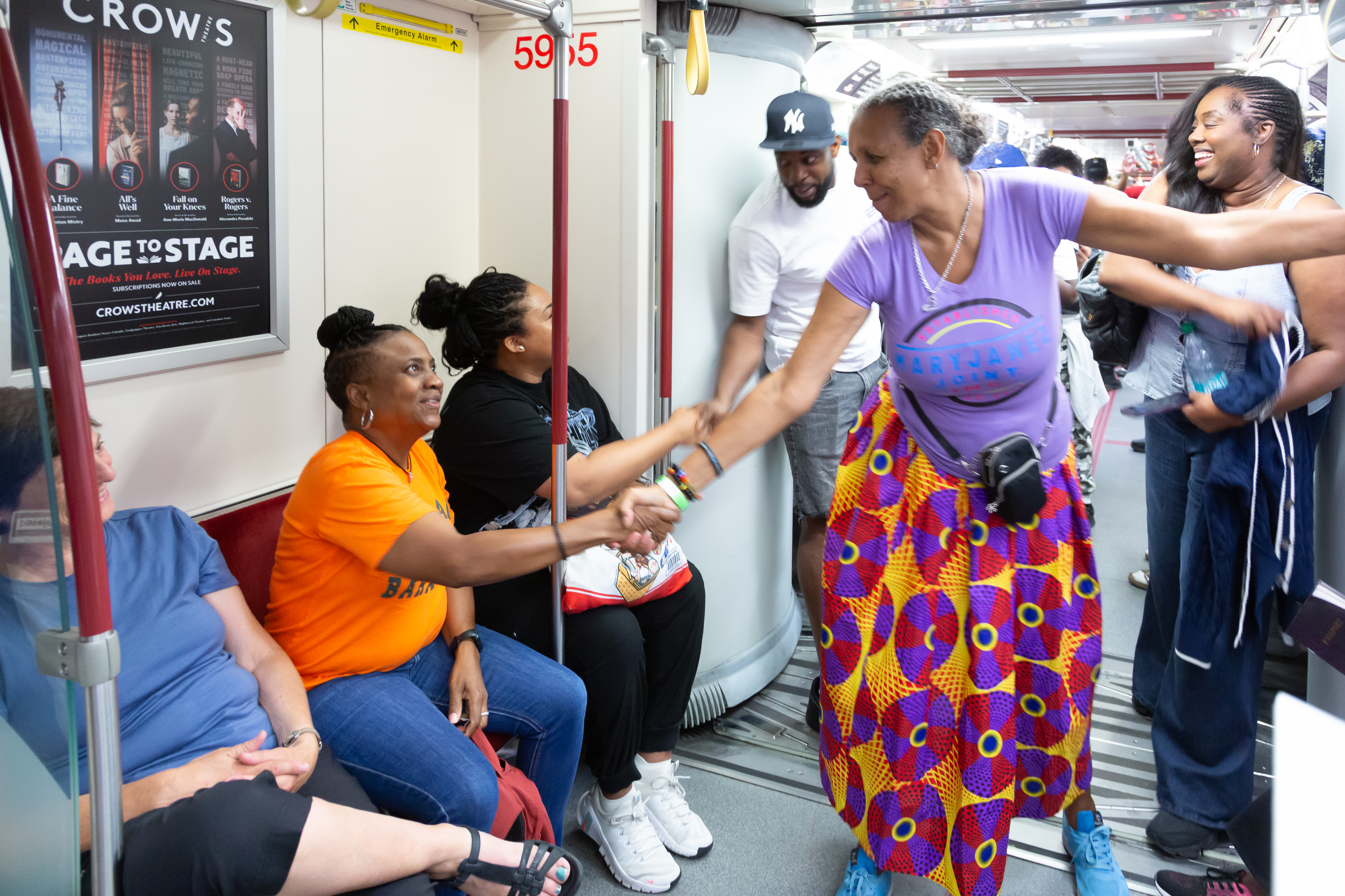 A group of people on a TTC subway train. One person in a purple shirt is standing and holding hands with a person in an orange shirt sitting down. They are both smiling.