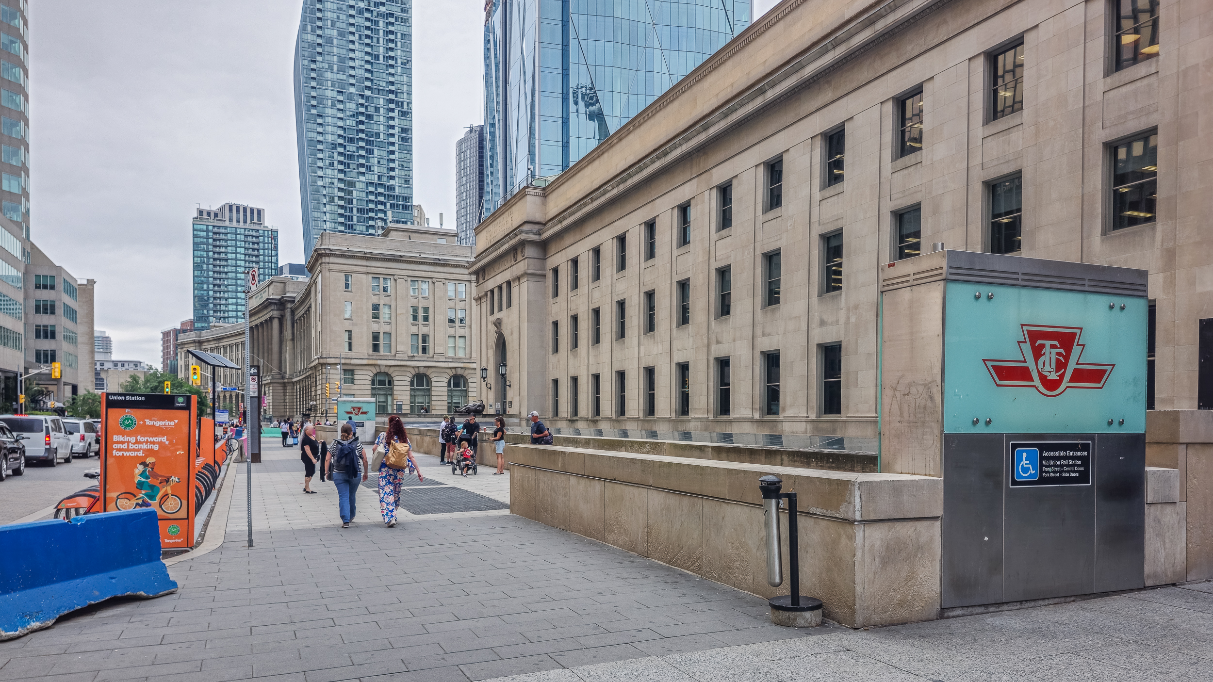Exterior view of Union Station, with a teal TTC subway entrance sign, pedestrians walking along the sidewalk, and downtown Toronto towers in the background.