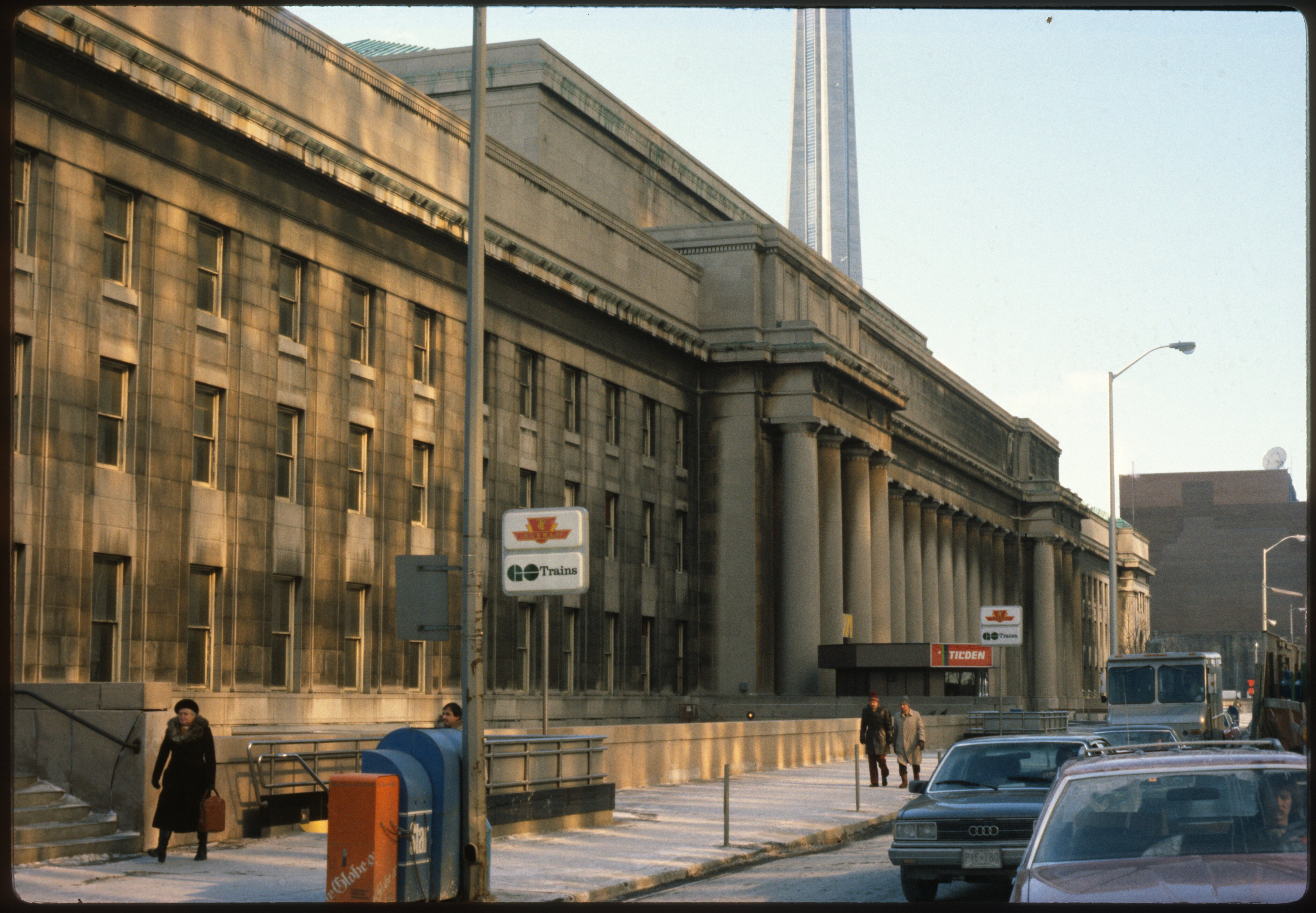 Vintage photo of Union Station’s exterior with a TTC and Go Trains sign, pedestrians on the sidewalk, vintage cars parked on the street, with the CN Tower in the background