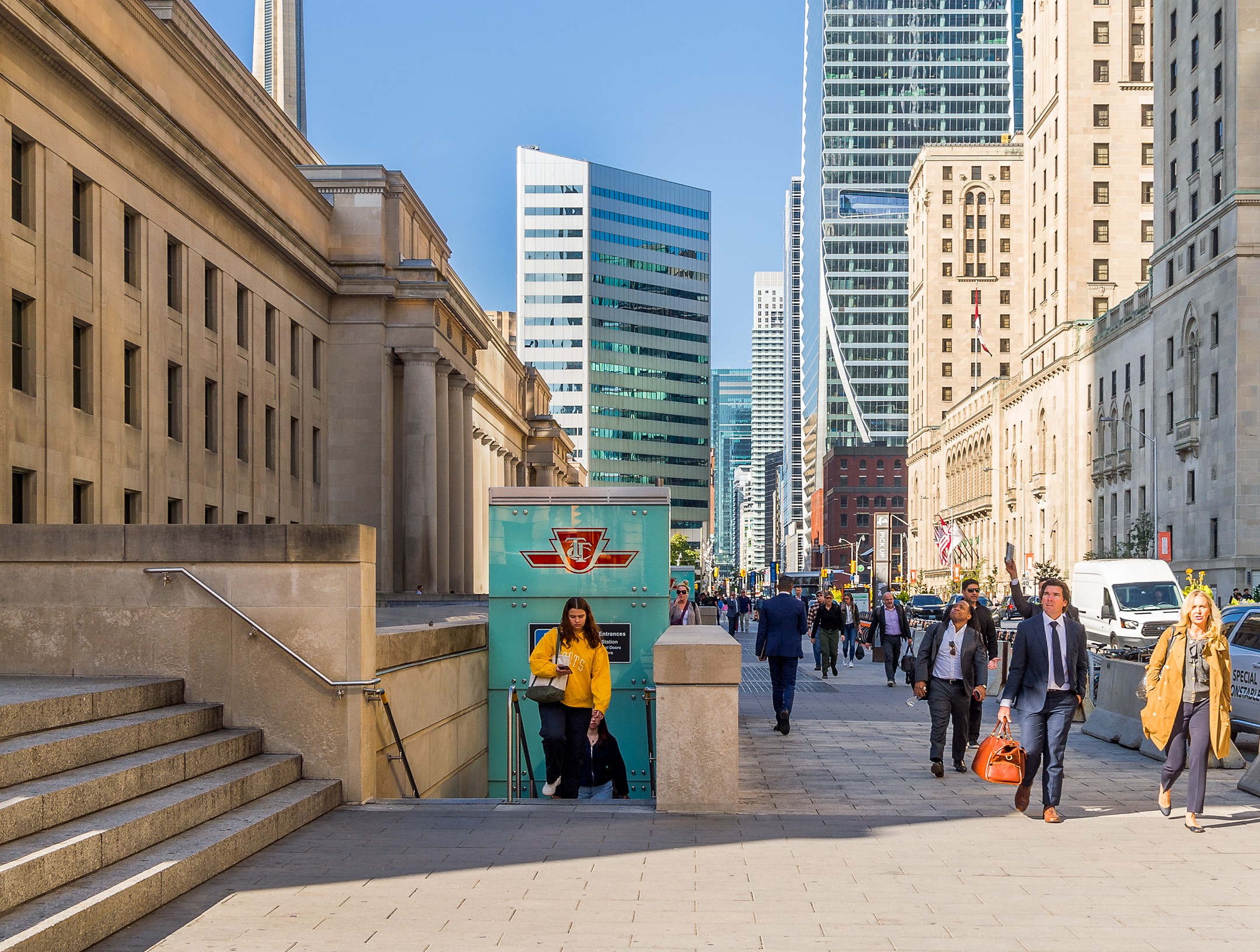 Modern photo of Front Street outside Union Station, with a TTC entrance, pedestrians, vehicles, the CN Tower and other skyscrapers in the background