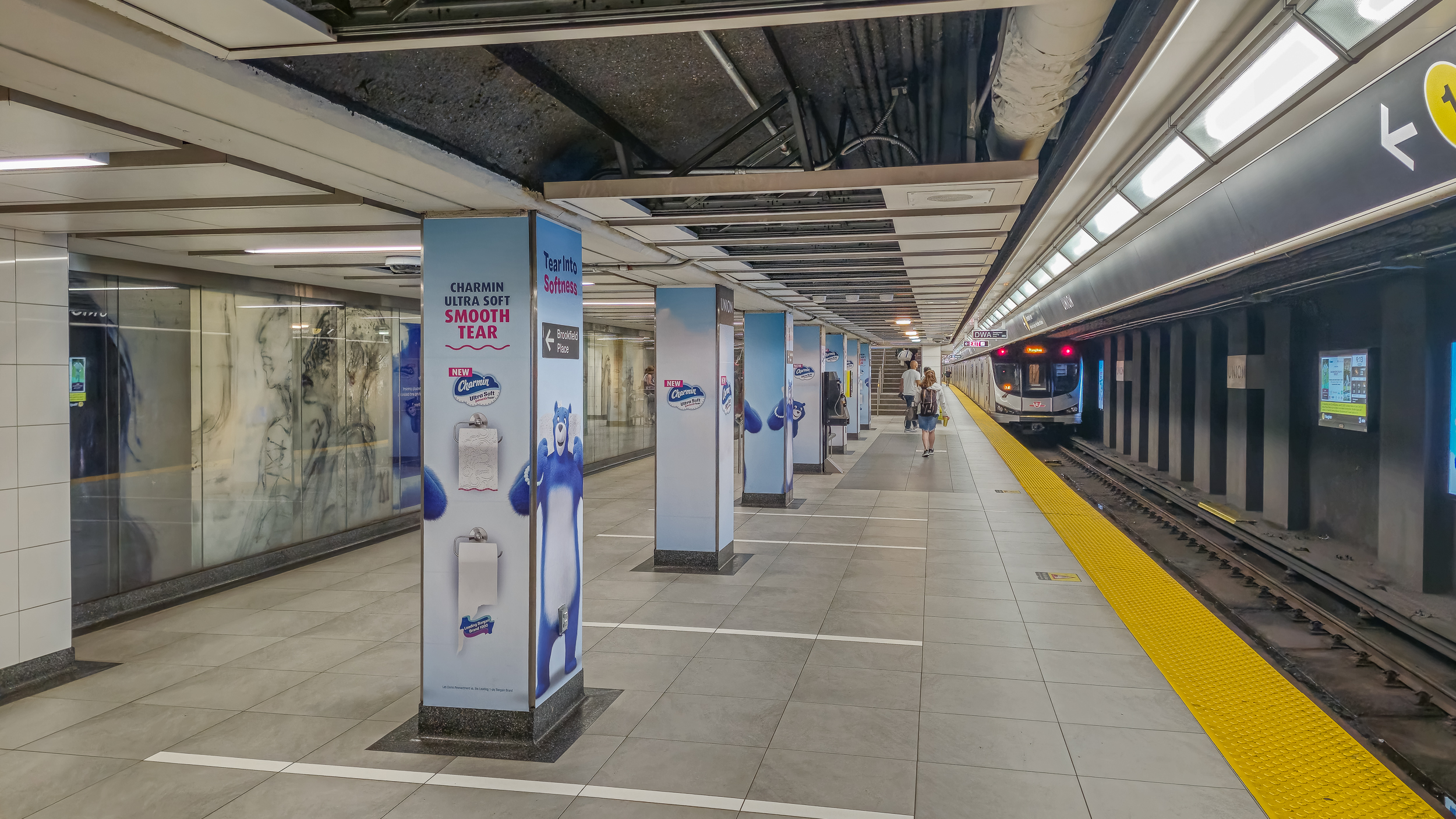 Subway platform with a train arriving, artwork, columns in the background with passengers on the platform