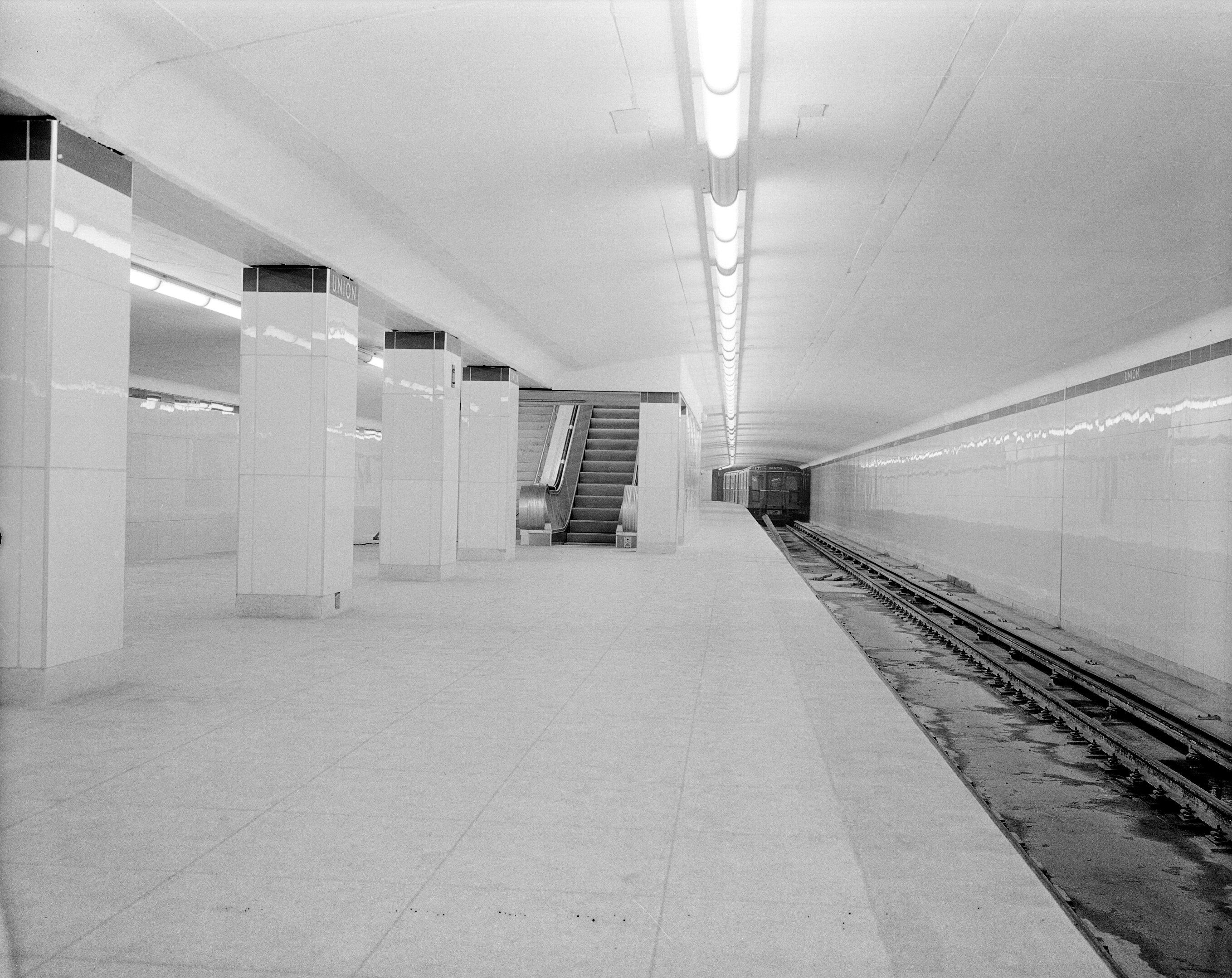 Black-and-white photo of TTC’s Union Station subway platform, with columns, stairs, and a train approaching on the tracks.