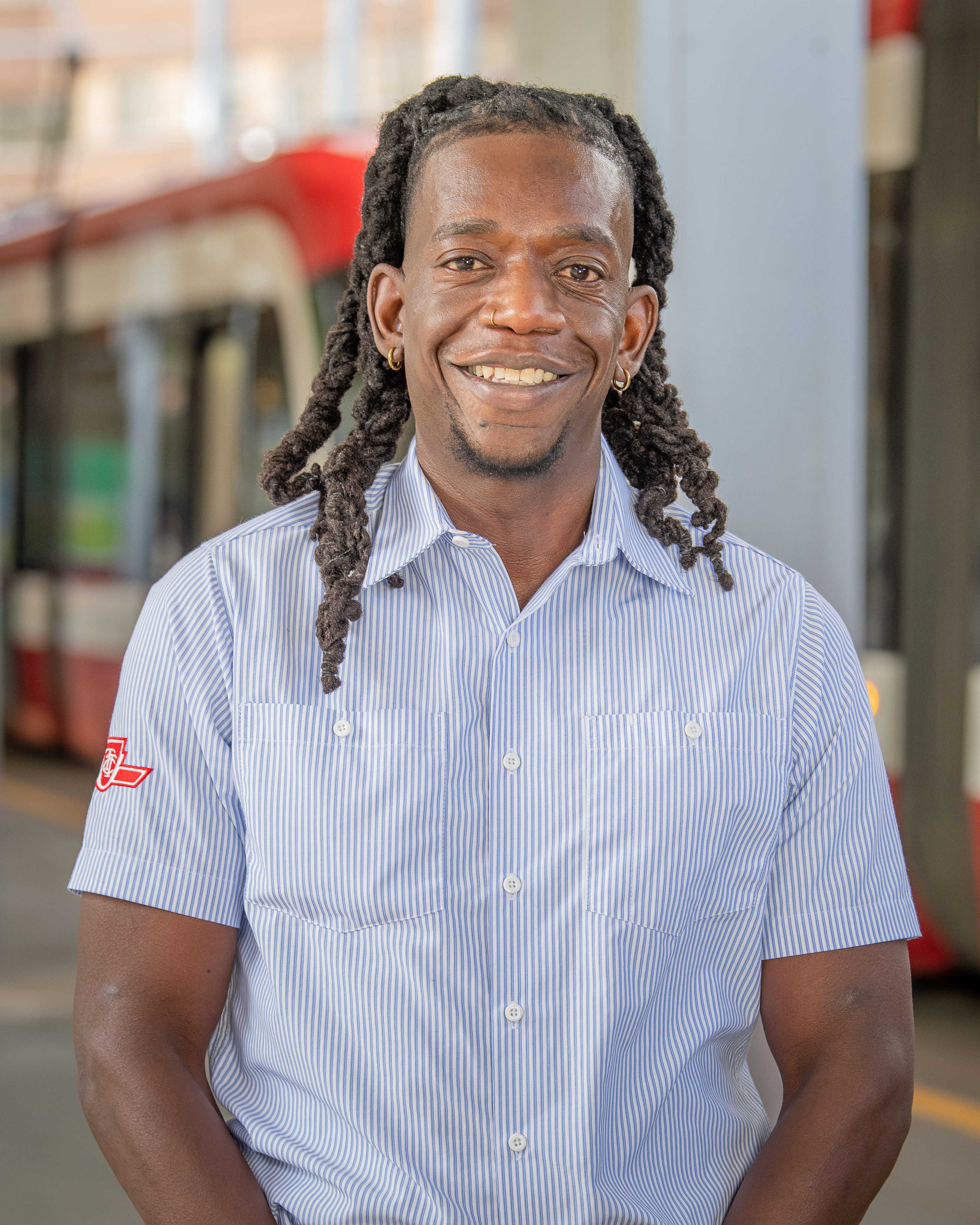 A TTC light rail transit operator smiling in a blue and white striped short-sleeve button up shirt with the TTC logo on their arm.