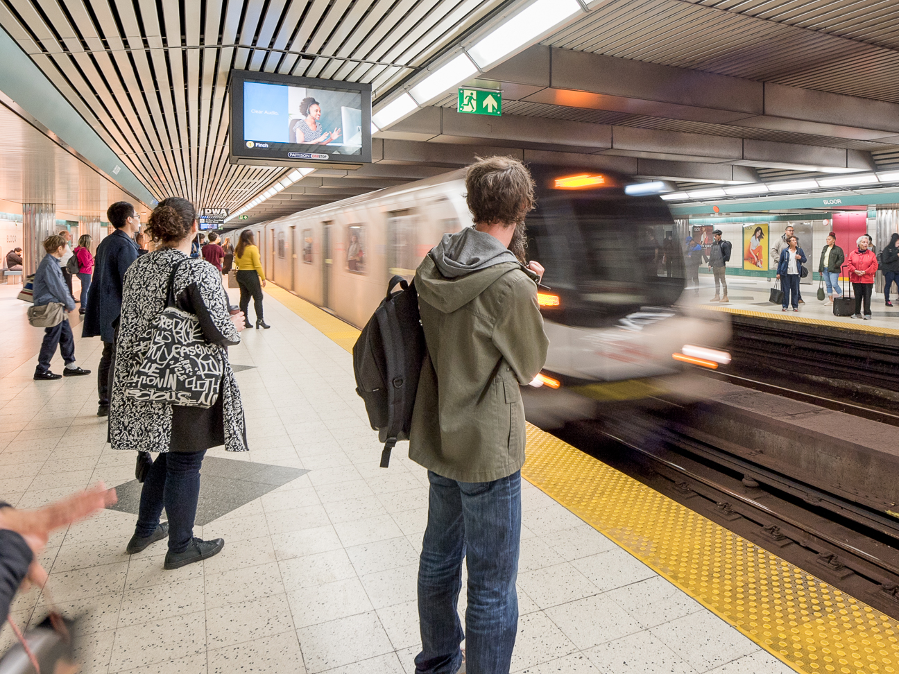 People standing on a subway platform, including one person wearing a backpack, as a TTC subway train pulls into the station.