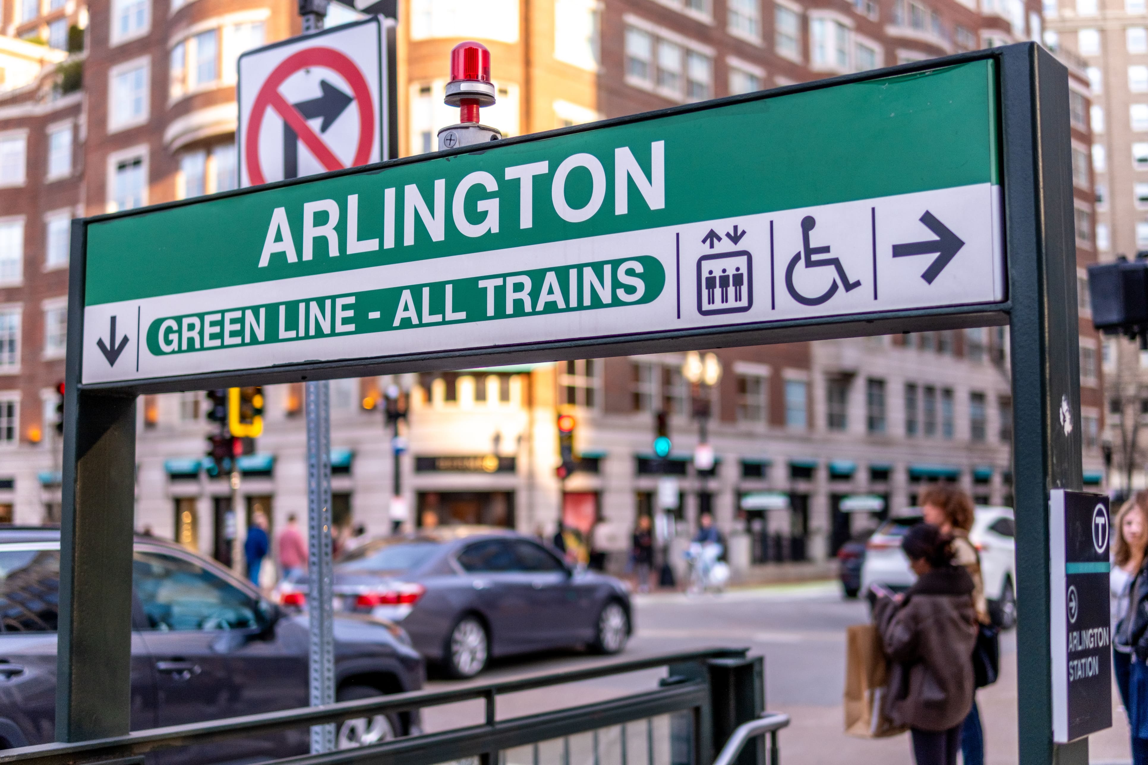 A green and white entrance sign to Boston's Arlington Station Green Line with people and cars in the background.