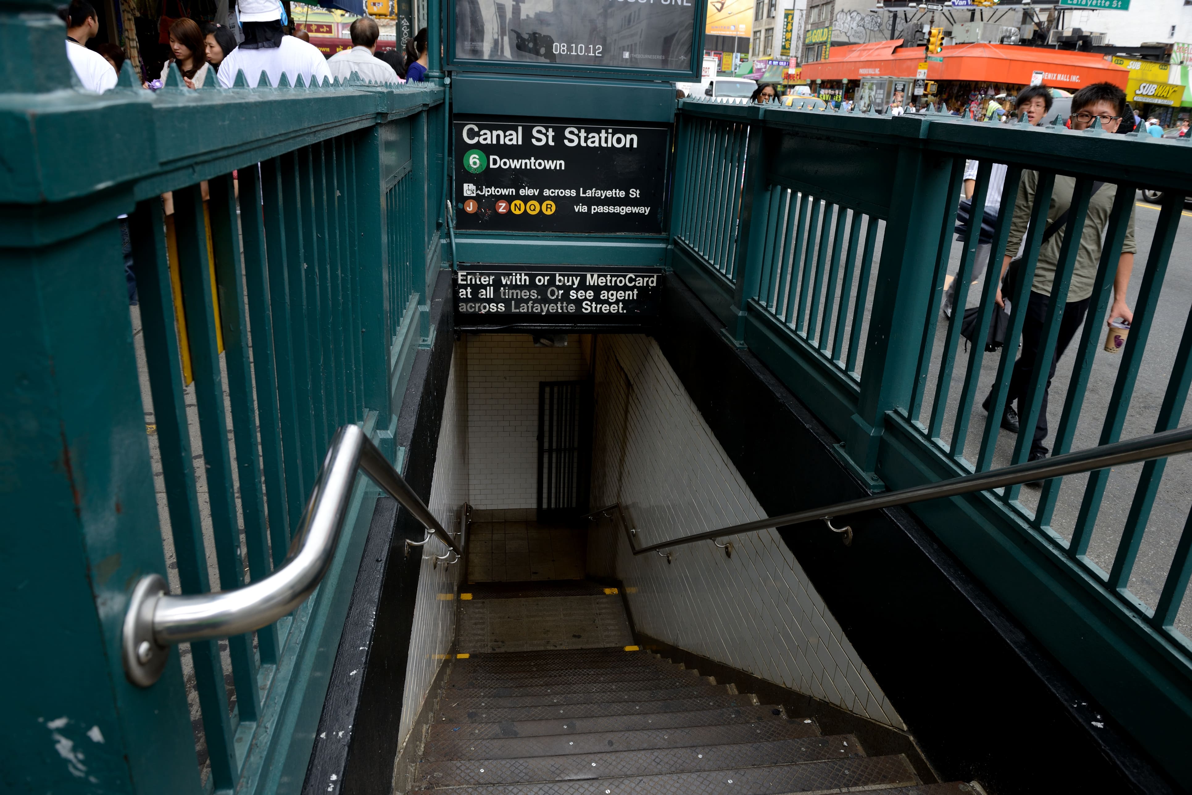 A stairwell on the street with green railings leading down to New York City's Canal Street Station and pedestrians on either side of the entrance.