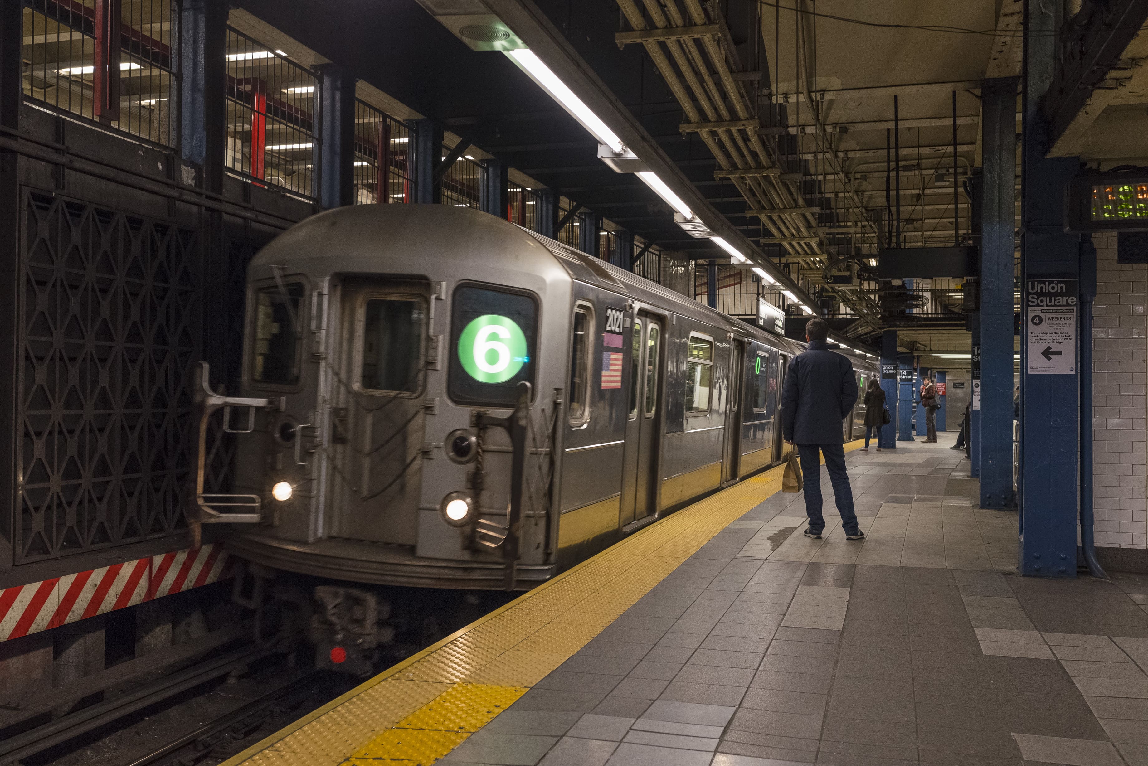 A New York City 6 Train arriving at a station platform where customers are waiting to board.