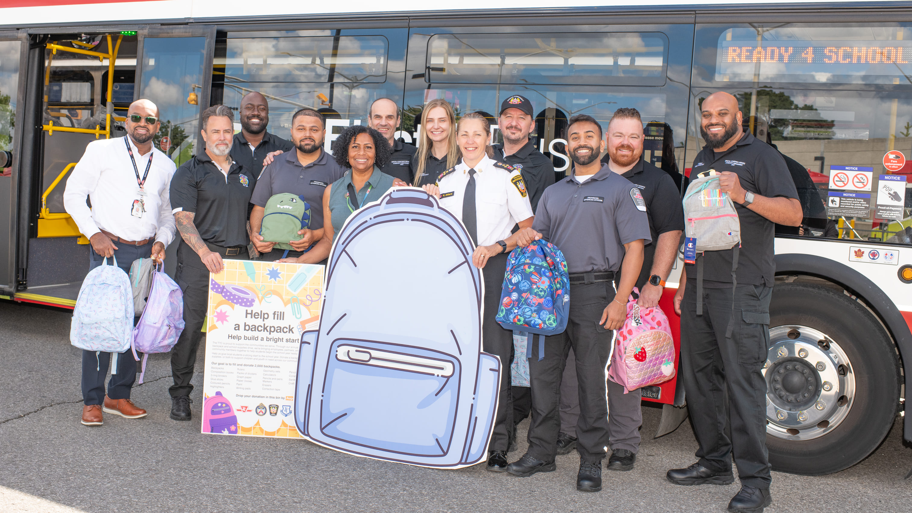 Group photo of TTC staff and special constables holding donated backpacks in front of a parked TTC Backpack Drive bus