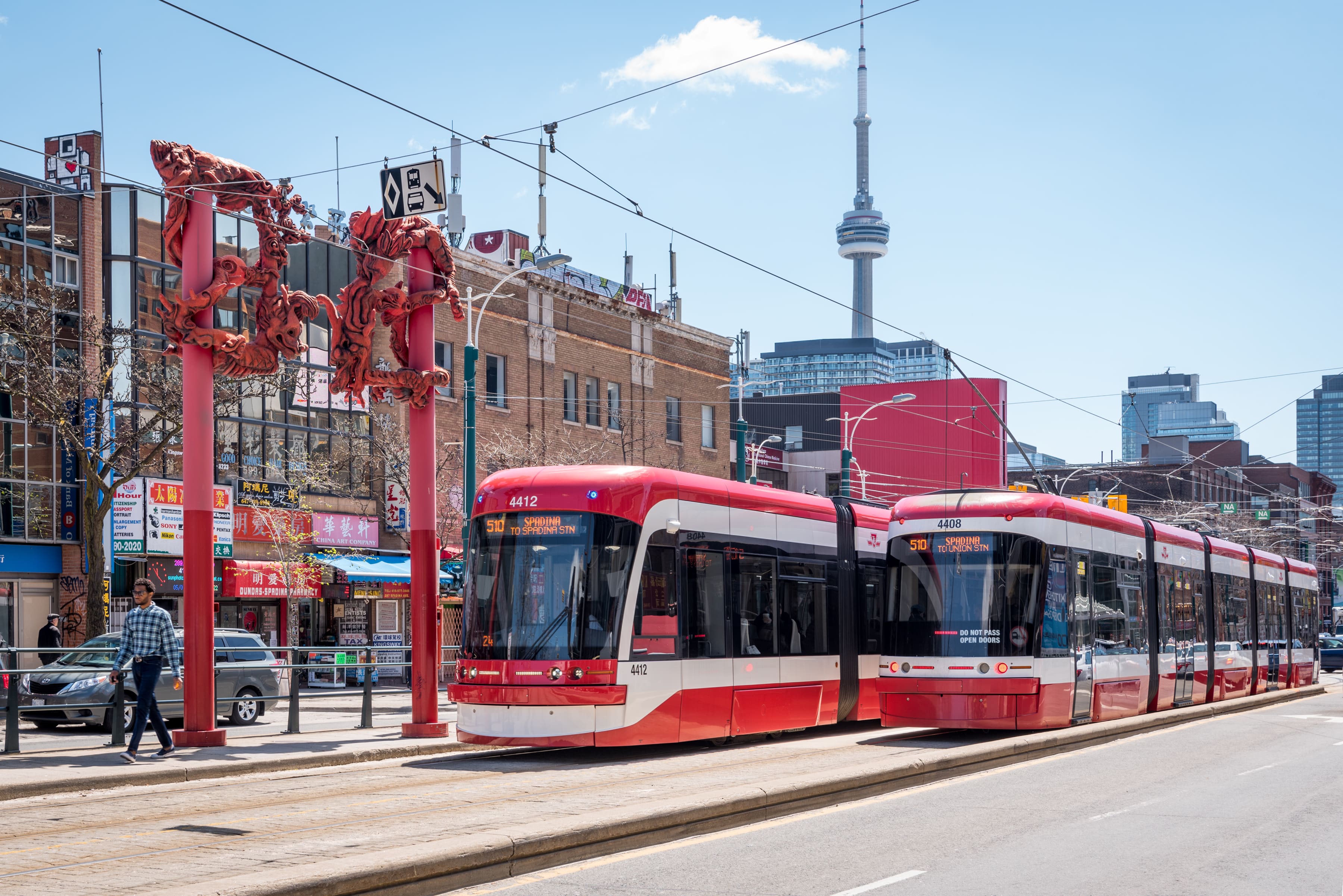 Two red and white TTC streetcars passing each other beside a red gateway structure in Chinatown. Buildings and the CN Tower are visible in the background.