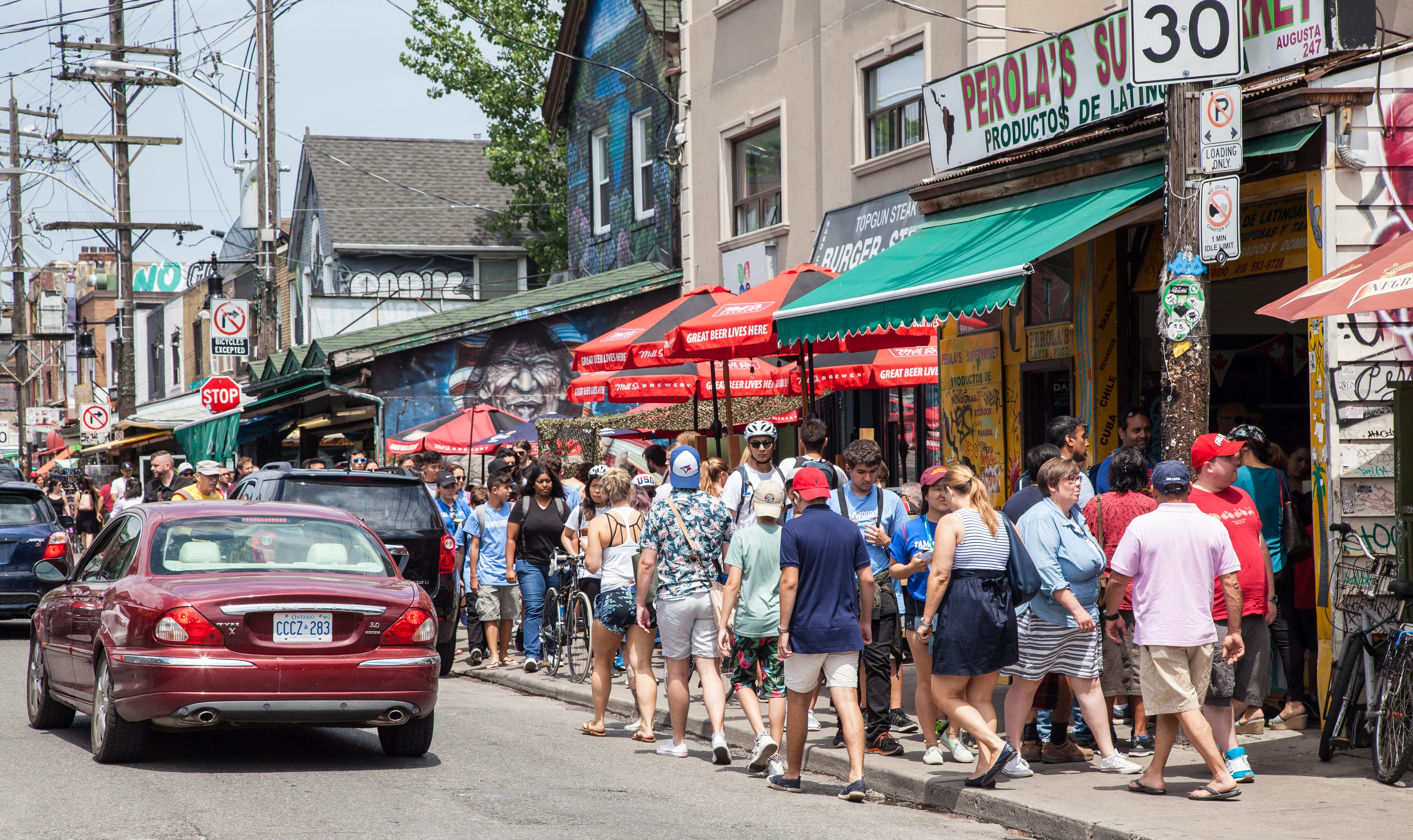 Toronto's Kensington Market in the summer with crowds of people on the sidewalk in front of shops and cars on the street.