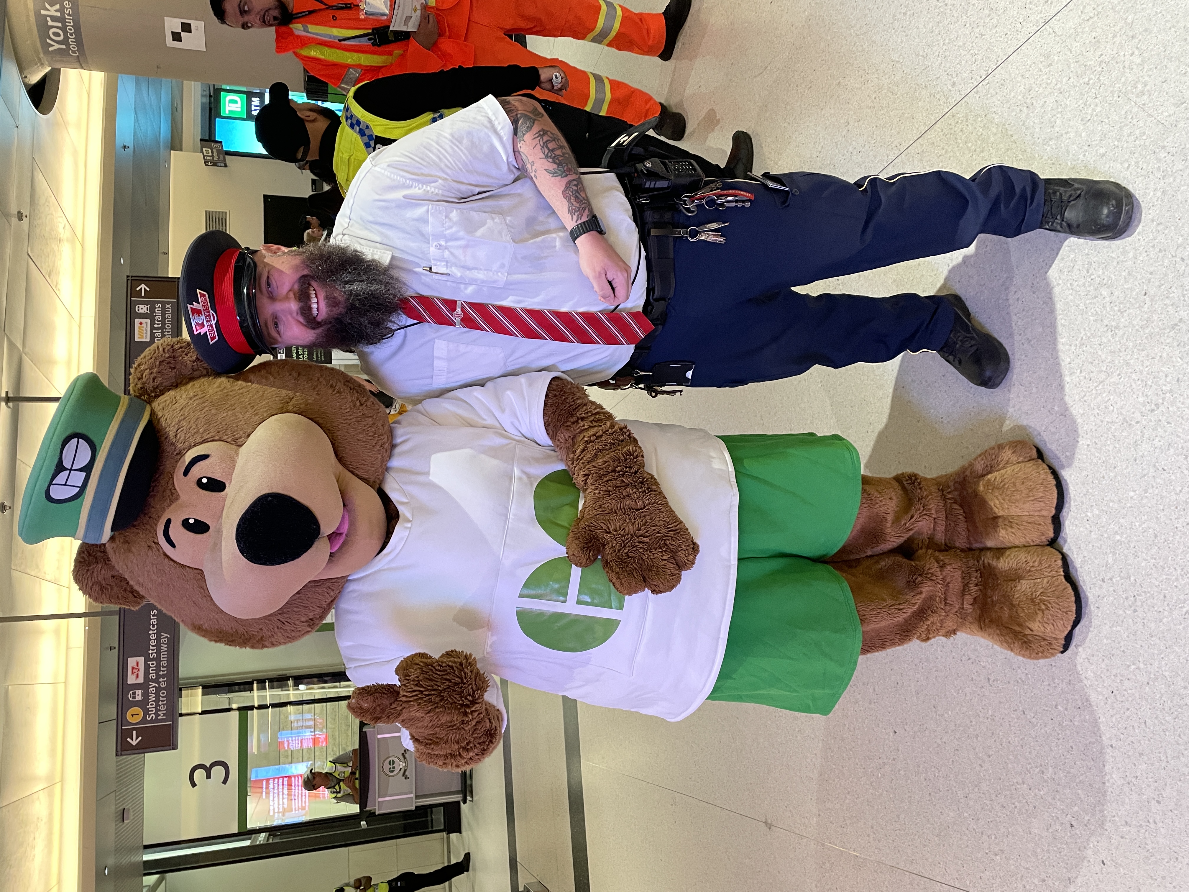  A TTC Mobile Supervisor in uniform standing beside a person wearing a GO Transit bear mascot costume