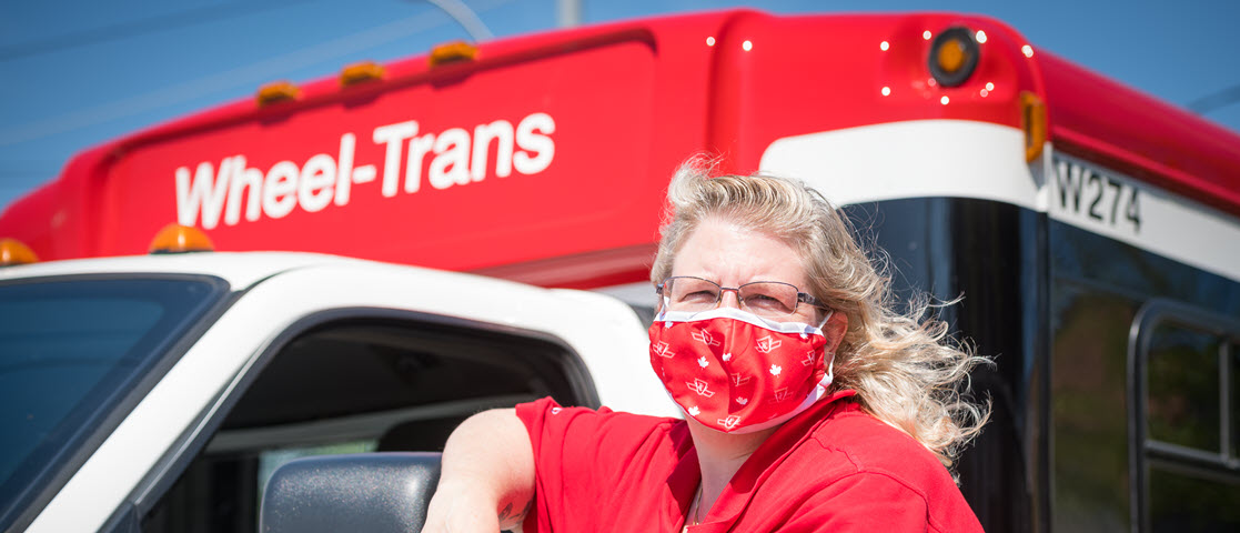 TTC Wheel-Trans operator wearing a TTC face mask posed for a photo beside her Wheel-Trans vehicle on a sunny day.