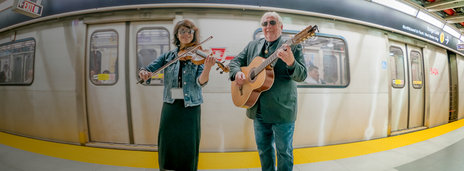 Image of a female musician with violin and male musician with guitar in front of a subway train